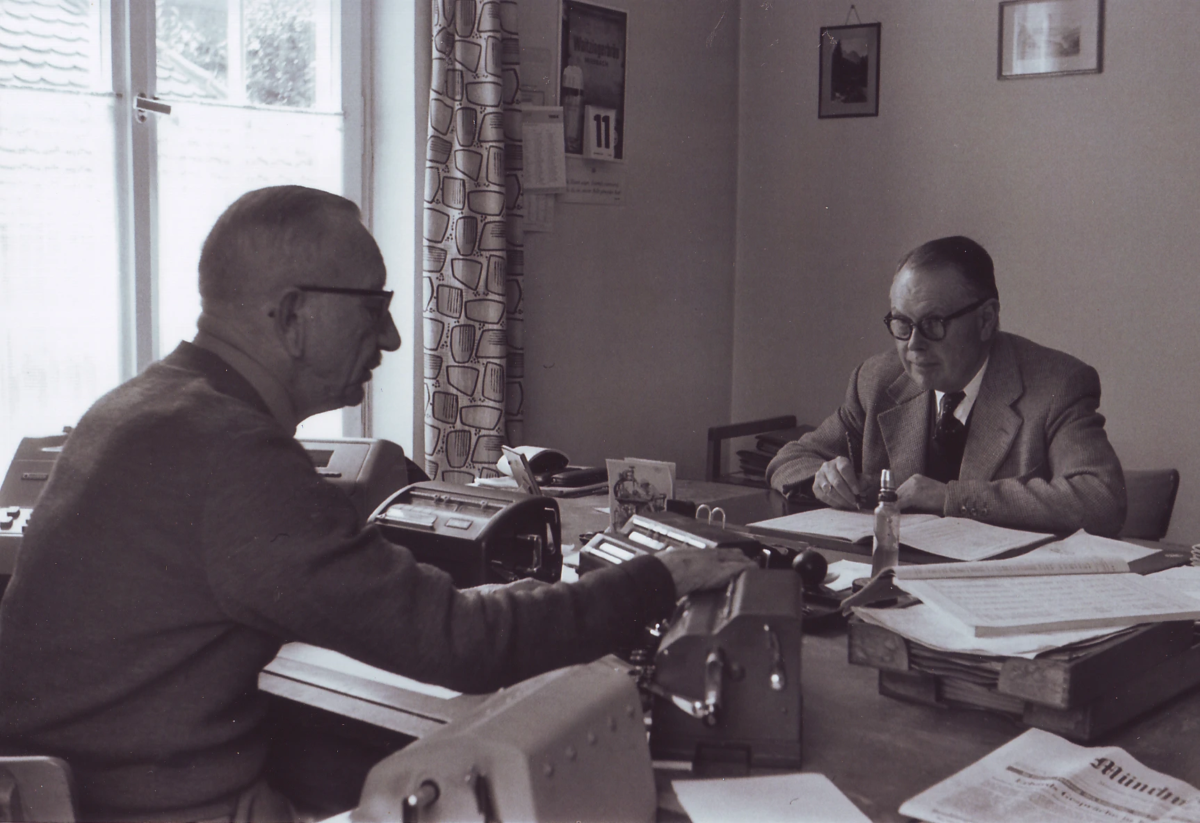 Two men in front of machines and papers at desk